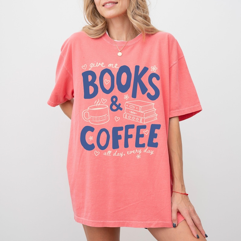 May include: A coral-colored short-sleeve t-shirt featuring the blue text "give me BOOKS & COFFEE" and "all day, every day". The design includes white line art of a coffee mug and a stack of three books, accented with small hearts and flowers.