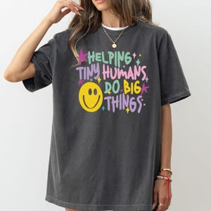Peut inclure: Un t-shirt gris anthracite à manches courtes avec une impression graphique multicolore portant l'inscription "HELPING TINY HUMANS DO BIG THINGS" en lettres vertes, roses et violettes, accompagnée d'un grand smiley jaune et de petites étoiles.
