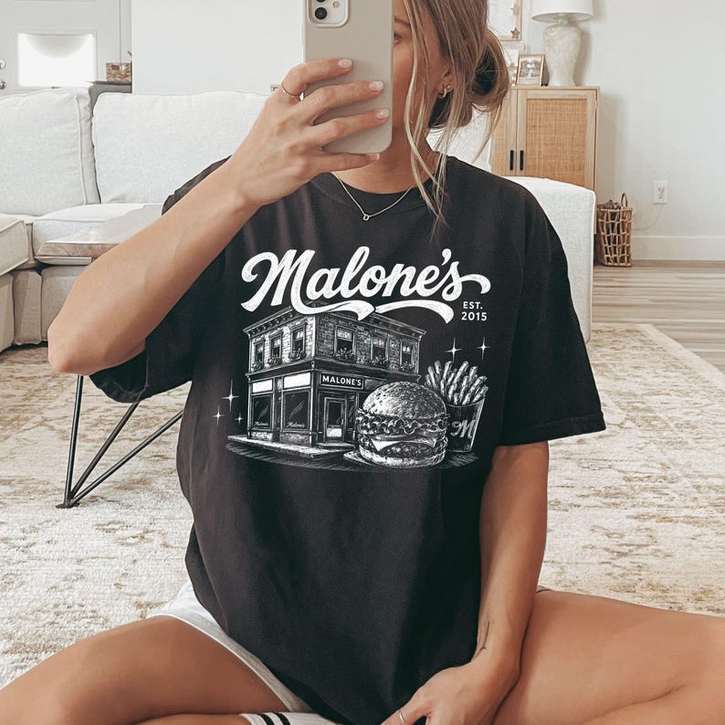 K&ouml;nnte beinhalten: Schwarzes T-Shirt mit dem Text "Malone's" in einer stilisierten Schriftart &uuml;ber einer detaillierten Illustration eines Restaurants, eines Burgers und Pommes Frites. Das Shirt enth&auml;lt auch den Text "EST. 2015".