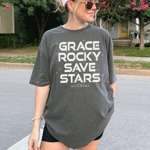May include: Dark grey t-shirt with the words "GRACE ROCKY SAVE STARS" in large white block letters. The text "PROJECT HAILMARY" is printed below. The shirt has short sleeves and a relaxed fit.