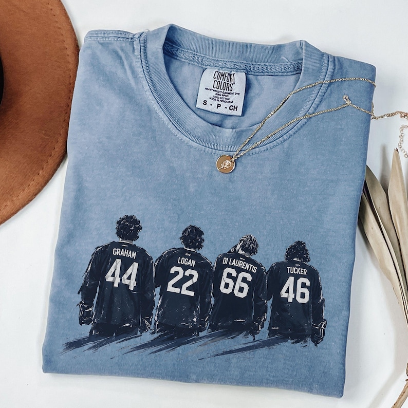 May include: A blue jean Comfort Colors t-shirt featuring a graphic of four hockey players with their jersey numbers and names