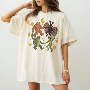 May include: An off-white short-sleeve t-shirt featuring a graphic print of a brown Bigfoot, a black moth-like creature, a green frog, and a green alien holding hands in a circle, surrounded by yellow stars and a crescent moon.