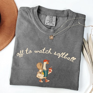 May include: Gray Comfort Colors t-shirt with the phrase "off to watch softball" in a cursive font. The shirt features a cartoon goose wearing a baseball uniform, holding a drink and a bag. A silver necklace with a pendant is also visible.