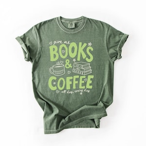 May include: A sage green short-sleeve t-shirt featuring a light green graphic print that reads "give me BOOKS & COFFEE all day, every day" alongside illustrations of a coffee mug and a stack of three books, accented with small hearts and flowers.
