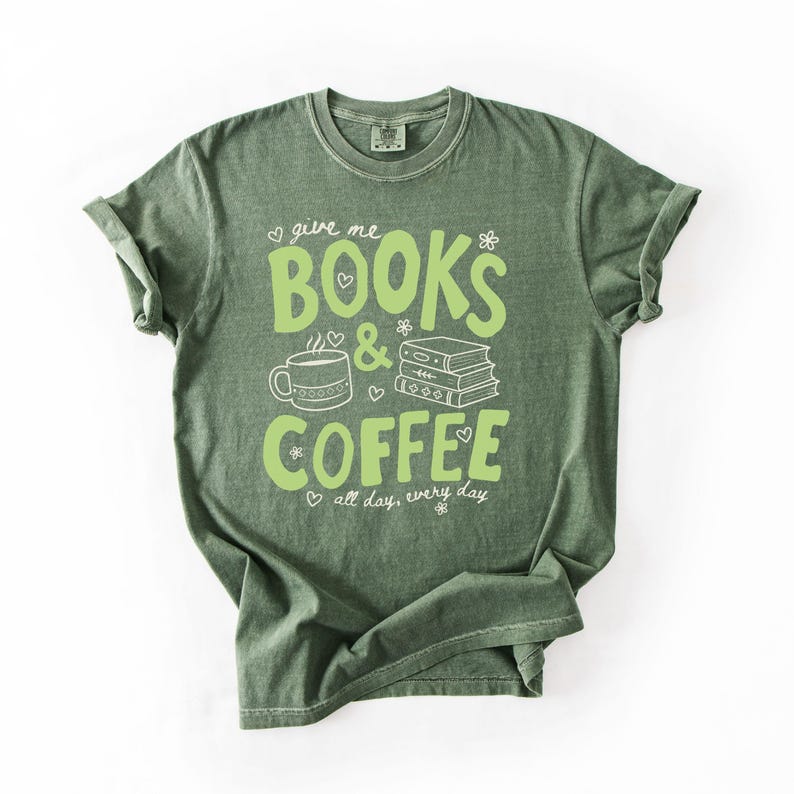 May include: A sage green short-sleeve t-shirt featuring a light green graphic print that reads "give me BOOKS & COFFEE all day, every day" alongside illustrations of a coffee mug and a stack of three books, accented with small hearts and flowers.