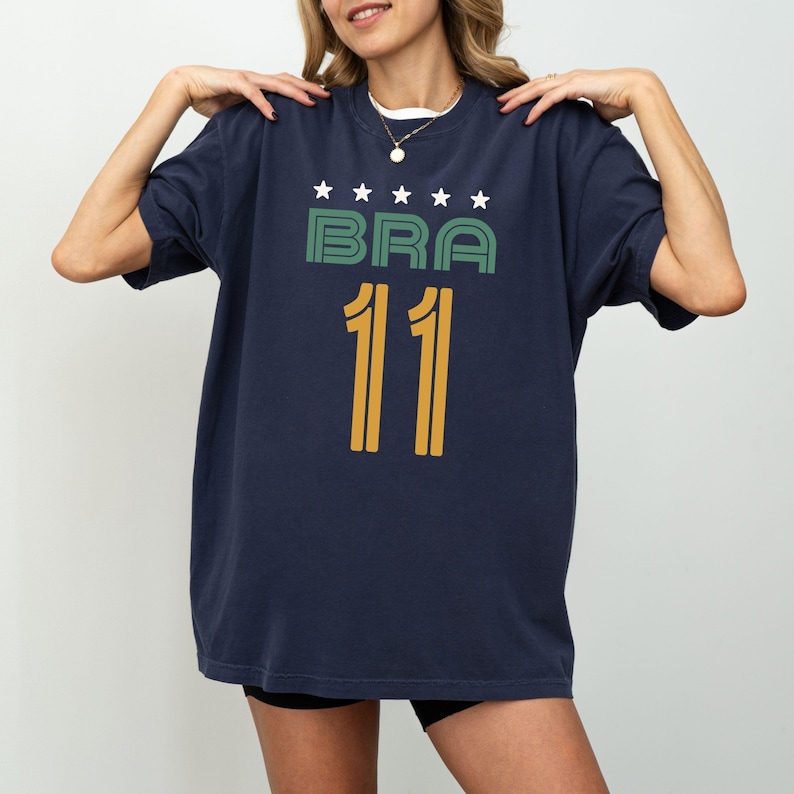 Peut inclure: T-shirt bleu marine avec le mot "BRA" en vert, surmont&eacute; d'&eacute;toiles blanches, et le num&eacute;ro "11" en jaune. Le t-shirt est de style d&eacute;contract&eacute;.