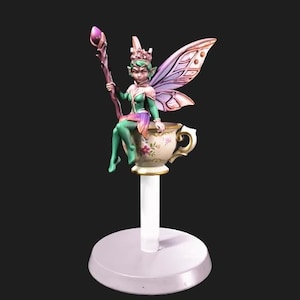 May include: A miniature figurine of a green-skinned fairy with large purple-tinted wings, wearing a crown and holding a staff with a purple orb, sitting on a floral-patterned teacup that is mounted on a white cylindrical post and circular base.