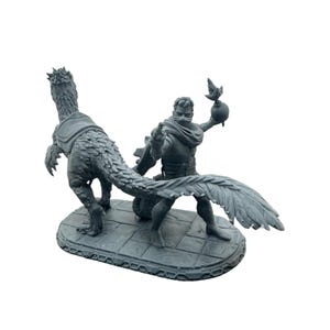 Op de afbeelding: Een grijze beeldje dat een fantasie scène uitbeeldt. Een figuur met een sjaal en een wapen staat tegenover een gevederd dinosaurusachtig wezen. De basis is een steenachtig oppervlak.