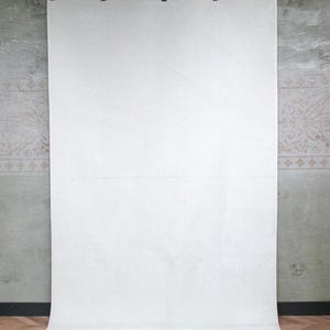 May include: A large, rectangular white textile hanging vertically from a black rod with five metal clips. The fabric features a subtle, repeating geometric pattern across its surface and has a fringed edge along the bottom hem.