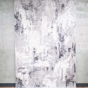 May include: A rectangular area rug featuring an abstract, distressed pattern in shades of white, light grey, and charcoal grey. The design displays a weathered, textured appearance with uneven colour distribution across the entire surface of the rug.