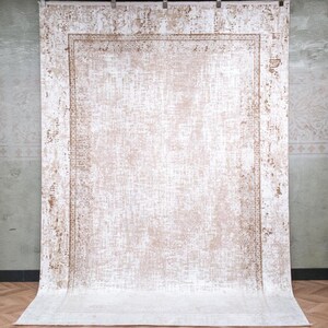 May include: A rectangular area rug featuring a distressed, faded pattern in shades of beige and white. The design includes a central field with a textured, weathered appearance, surrounded by a decorative border with intricate, repeating geometric motifs.