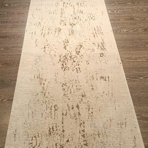 May include: A long, rectangular runner rug featuring a cream-coloured base with a distressed, weathered pattern in shades of light brown and metallic gold. The rug has a textured surface and short, frayed fringe detailing along the narrow ends.