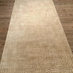 May include: A long, rectangular runner rug featuring a light beige and tan colour palette. The surface displays a repeating geometric pattern of interconnected, rounded lines and parallel stripes that create a continuous, maze-like visual effect.