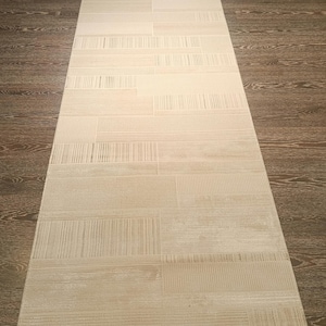 May include: A long, rectangular runner rug featuring a neutral beige and cream colour palette. The surface displays a repeating geometric pattern composed of alternating blocks of horizontal lines and solid textures, creating a subtle, layered effect.