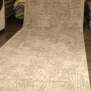 May include: A long rectangular runner rug featuring a light beige and cream colour palette. The surface displays a repeating geometric pattern composed of interconnected lines, curves, and rectangular blocks that create a textured visual effect.