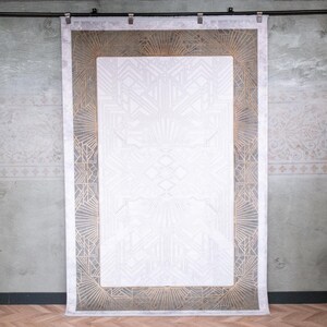 May include: A rectangular wall hanging featuring a white centre panel with subtle geometric line patterns. The border displays a dark grey background with gold-coloured sunburst and linear motifs. The piece is suspended from a black metal rod.