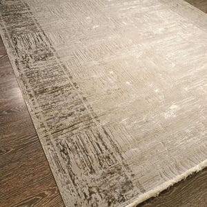 May include: A rectangular area rug featuring a light grey and silver colour palette with a distressed, textured pattern. The design includes a darker grey border with a weathered, scratch-like motif and a fringe trim along the short edge.