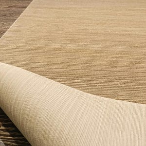 May include: A rectangular area rug with a textured, ribbed surface in a neutral tan colour. The rug is partially folded over, revealing a lighter, off-white woven backing with a distinct linear grid pattern on the underside of the material.