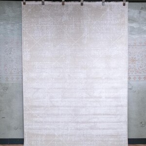 May include: A large rectangular area rug in a light beige and off-white colour palette. The rug features a subtle, distressed diamond pattern across its surface and is suspended from a black metal rod by six silver metal clips along the top edge.
