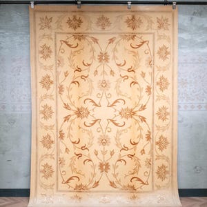 Könnte beinhalten: Ein rechteckiger Teppich mit beigem Hintergrund und einem symmetrischen Muster aus braunen Blumenmotiven und geschwungenen Ranken. Das Design umfasst eine breite Bordüre mit sich wiederholenden Blütenelementen und einer zentralen Anordnung.