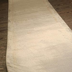 May include: A long, rectangular runner rug featuring a distressed, faded pattern in shades of cream, beige, and light grey. The surface has a textured, weathered appearance with subtle variations in colour density across the entire length.