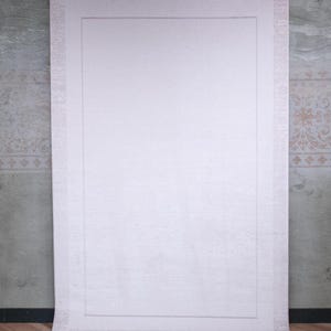 May include: A large, rectangular, off-white area rug featuring a solid central panel framed by a wide, textured border. The rug is suspended vertically from a black metal rod by four silver clips, displaying a uniform, light-coloured surface.