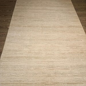 May include: A rectangular area rug featuring a textured, horizontal striped pattern in alternating shades of cream and light beige. The rug has a low-pile construction with a subtle, uneven weave and short, frayed fringe detailing along the top and bottom edges.