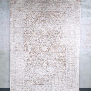 May include: A large rectangular area rug featuring a distressed, faded pattern in shades of cream, beige, and light brown. The design includes a central medallion surrounded by intricate floral and geometric motifs, all enclosed within a decorative border.