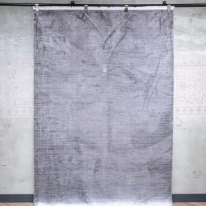 May include: A rectangular area rug featuring a solid light grey colour with subtle tonal variations and a soft, slightly reflective sheen. The rug has a short, dense pile texture and is finished with a clean, straight edge along the bottom border.