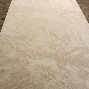 May include: A rectangular area rug featuring a cream and off-white colour palette. The surface displays a textured, abstract pattern composed of layered, brushstroke-like shapes arranged in a repeating, geometric, herringbone-inspired orientation.
