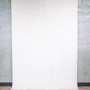 May include: A rectangular area rug featuring a cream-coloured surface with a repeating pattern of raised, textured lines forming concentric semi-circles and straight geometric segments. The rug is suspended from a black rod by metal clips.
