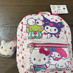 May include: White mini backpack with a pink zipper and a small matching keychain. The backpack features cartoon characters and red cherries on a white background. The keychain is a white Hello Kitty face with a pink bow.