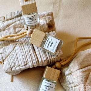 May include: Three small glass oil diffusers with wooden caps and labels. The labels read "White Orris & Sandalwood", "Bohemian Rose", and "Lily of the Valley". Each diffuser has a tan cord and wooden bead for hanging. The background is a neutral beige.