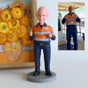 Può includere: Una statuetta bobblehead personalizzata di un uomo in abiti da lavoro, che fa un gesto di pollice in su. La figura indossa una camicia da lavoro a righe arancioni e blu, pantaloni blu scuro e scarpe nere. La statuetta è su una base circolare grigia. Un dipinto incorniciato di girasoli è sullo sfondo.