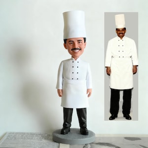 May include: A chef figurine wearing a tall white hat, white chef's coat with black buttons, and a white apron. The figurine has a smiling face and a mustache, with black trousers and shoes. The figurine is on a grey base.