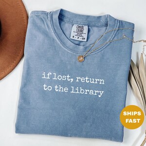 May include: A blue Comfort Colors t-shirt with the text "if lost, return to the library" in white. A gold necklace with a pendant rests on the shirt. The shirt is folded and has a small tag.