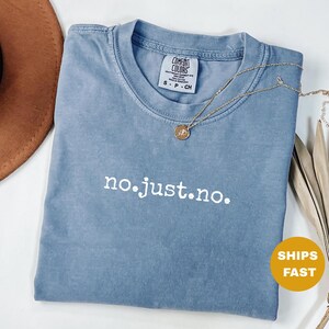 May include: A blue Comfort Colors t-shirt with the embroidered phrase "no.just.no." in white. A gold necklace with a pendant rests on the shirt. The shirt is folded and has a small tag. A brown hat is visible in the background.
