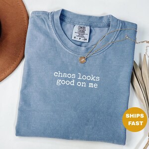 Könnte beinhalten: Ein blaues Comfort Colors T-Shirt mit dem weißen Schriftzug „chaos looks good on me“. Eine goldene Halskette mit Anhänger liegt auf dem Shirt. Ein brauner Hut ist in der oberen linken Ecke zu sehen.