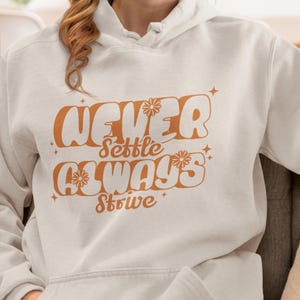 Puede incluir: Sudadera con capucha color crema con las palabras "NEVER Settle ALWAYS Strive" en naranja, con letras de estilo retro. El diseño incluye detalles florales y estrellas. La sudadera tiene un bolsillo delantero y una capucha con cordón.