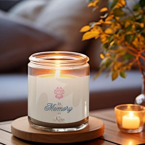 Puede incluir: Una vela encendida en un tarro de cristal con una etiqueta blanca con una huella de pata rosa y las palabras "In Memory of Kira". La vela está sobre un posavasos de madera, con una vela más pequeña en un votivo de cristal a la derecha. Una planta está en el fondo.