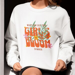 Puede incluir: Sudadera blanca con el texto "WAIT AND SEE THE GIRLS BLOOM SUMMER" en naranja, verde y rosa. El diseño incluye elementos florales y de hojas. La sudadera es de estilo casual y cómodo.