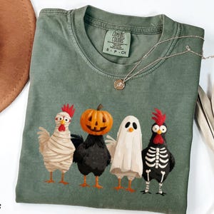 Puede incluir: Una camiseta de manga corta color verde musgo con un gráfico de cuatro pollos con disfraces de Halloween: una momia, un pollo con cabeza de calabaza, un fantasma y un esqueleto. La camiseta tiene cuello redondo y etiqueta Comfort Colors.