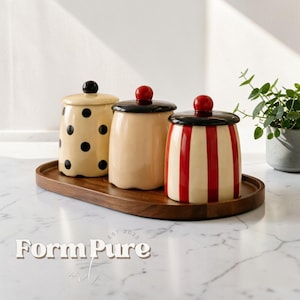 Puede incluir: Tres botes de cerámica con tapas sobre una bandeja de madera. Uno es de color crema con lunares negros, otro es crema liso y el tercero tiene rayas rojas y crema. Cada uno tiene una tapa negra y un pomo rojo o negro.
