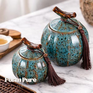 May include: Two ceramic jars with lids. The jars are teal with a speckled pattern and brown accents. Each jar has a brown handle on top and a long brown tassel. The jars are on a marble surface.