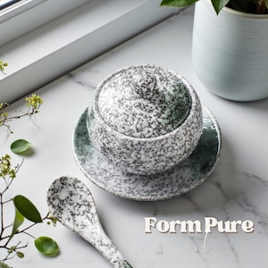 以下が含まれることがあります： 蓋、受け皿、スプーン付きのセラミック製サービングボウル。グレーとグリーンの色合いの斑点模様が特徴です。「Form Pure」の文字が右下に表示されています。ボウルは大理石の表面にあります。
