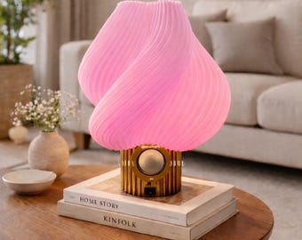 May include: A pink, sculptural table lamp with a swirling, pleated shade. The lamp sits on a gold-colored base with a circular speaker. The lamp is on a wooden coffee table with two books, "Home Story" and "Kinfolk".