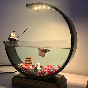 Puede incluir: Un acuario de cristal en forma de media luna con marco negro e iluminación LED integrada. Una figura en miniatura de una persona pescando se sienta en el borde. Dentro, un pez betta nada sobre grava de colores y un letrero que dice BALIK TUTMAK.