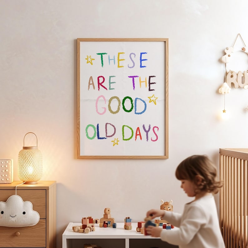 May include: A framed print with the words "THESE ARE THE GOOD OLD DAYS" in colorful, crayon-style lettering. The artwork is in a light wood frame, hanging on a white wall in a child's room. A child is playing with wooden toys in the foreground.