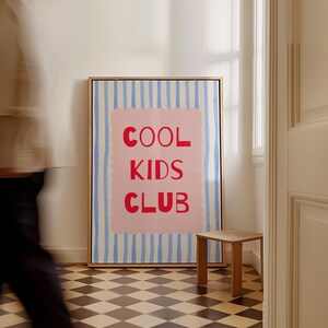 K&ouml;nnte beinhalten: Ein gerahmter Kunstdruck mit dem roten Schriftzug "COOL KIDS CLUB" auf rosa Hintergrund, vor einem Hintergrund aus blau-wei&szlig;en vertikalen Streifen. Das Kunstwerk befindet sich in einem Raum mit einem kleinen Holzschemel und einem Schachbrettboden.