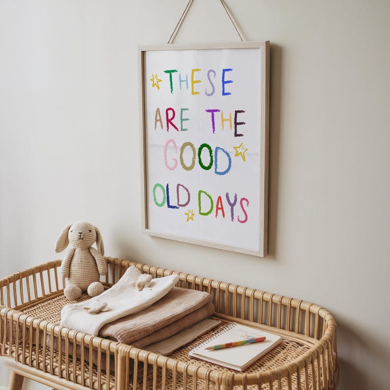 May include: A framed print with the words "THESE ARE THE GOOD OLD DAYS" in colorful crayon-style lettering. Below, a woven wicker bassinet holds folded blankets, a stuffed bunny, and a notebook with a rainbow pencil.